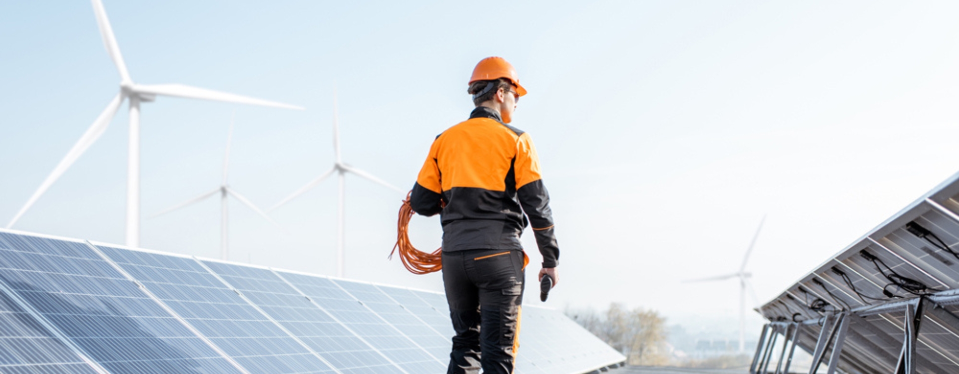 Man in orange hard hat walking past rows of solar panels with wind turbine in background